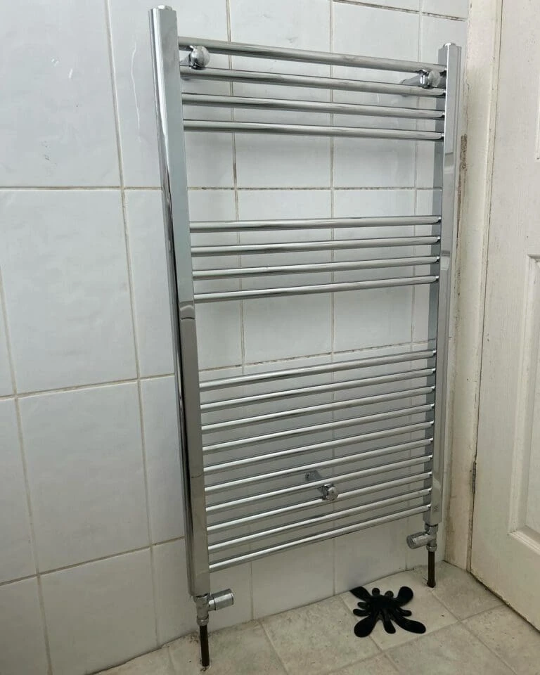 Chrome towel radiator mounted on tiled wall, featuring multiple horizontal bars and connected to plumbing. Black floor mat visible below.