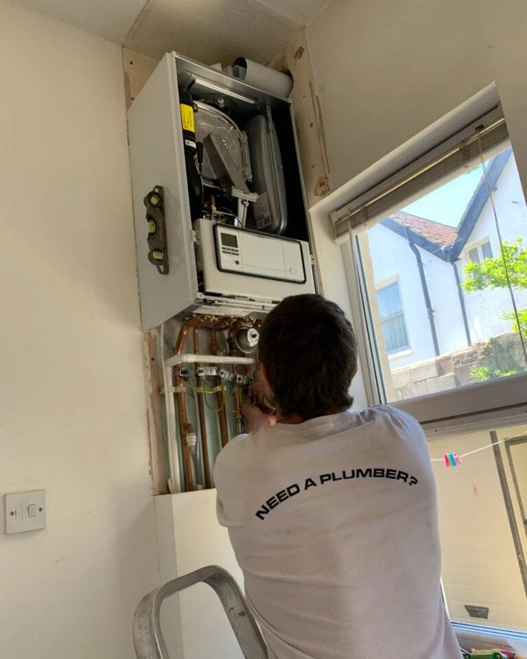 A person working on a wall-mounted boiler, wearing a t-shirt that says "NEED A PLUMBER?", on a ladder in a well-lit room.