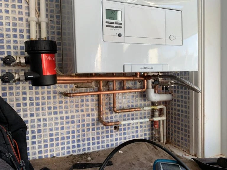 Copper piping and a boiler mounted on a tiled wall, showing plumbing connections and a filter with a gauge below.