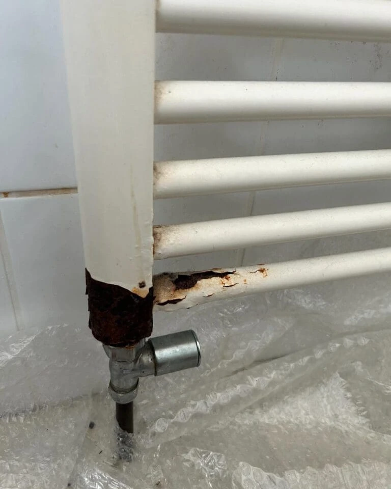 Rusty metal radiator pipe with corrosion at the base, adjacent to a silver valve and wrapped in bubble wrap.