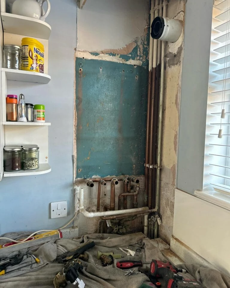 A kitchen corner showing exposed pipes, a blue wall with peeling paint, and tools on a covered surface beneath. Shelves hold jars and food items.