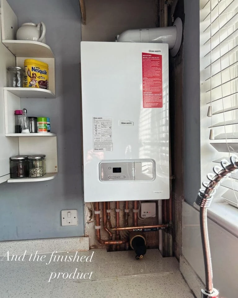 A wall-mounted boiler with exposed copper pipes, next to kitchen shelves containing jars and Nesquik, on a light-colored countertop.