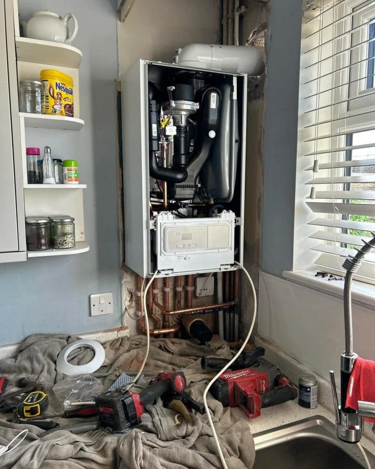 A kitchen with an open boiler cabinet, tools scattered on a countertop, and visible plumbing pipes; messy yet functional.