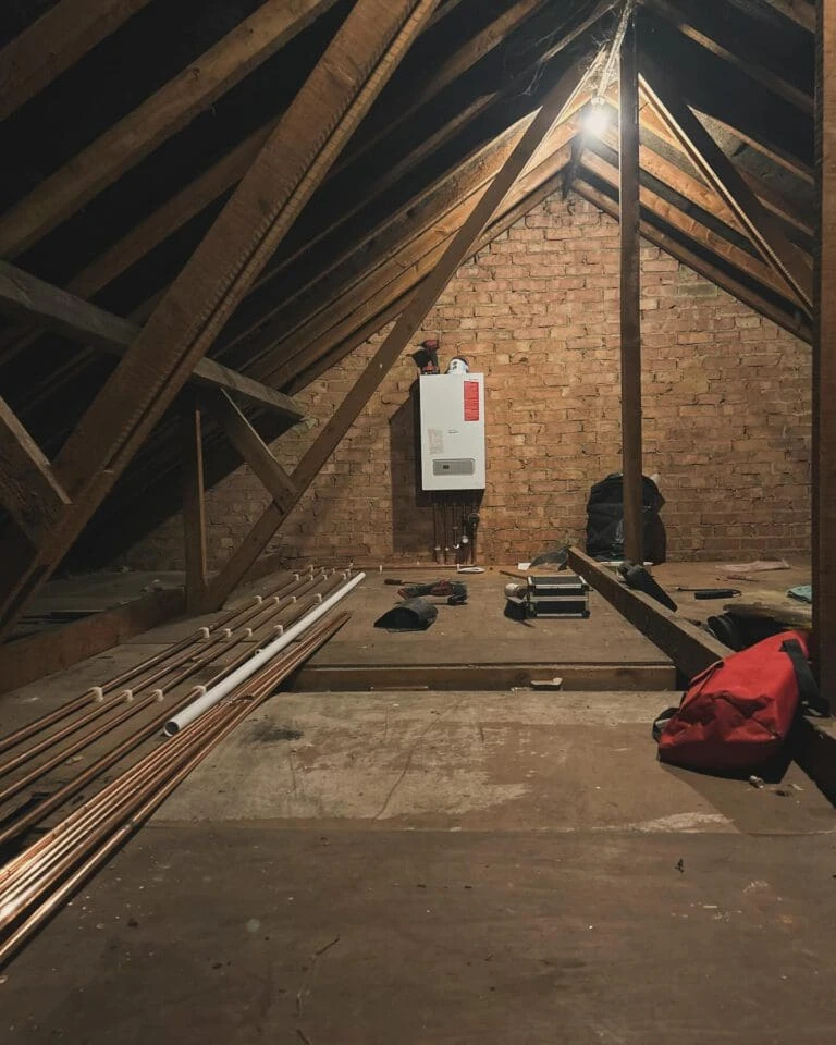 Dimly lit attic with wooden beams, brick walls, a boiler on the wall, and scattered tools and pipes on the floor.