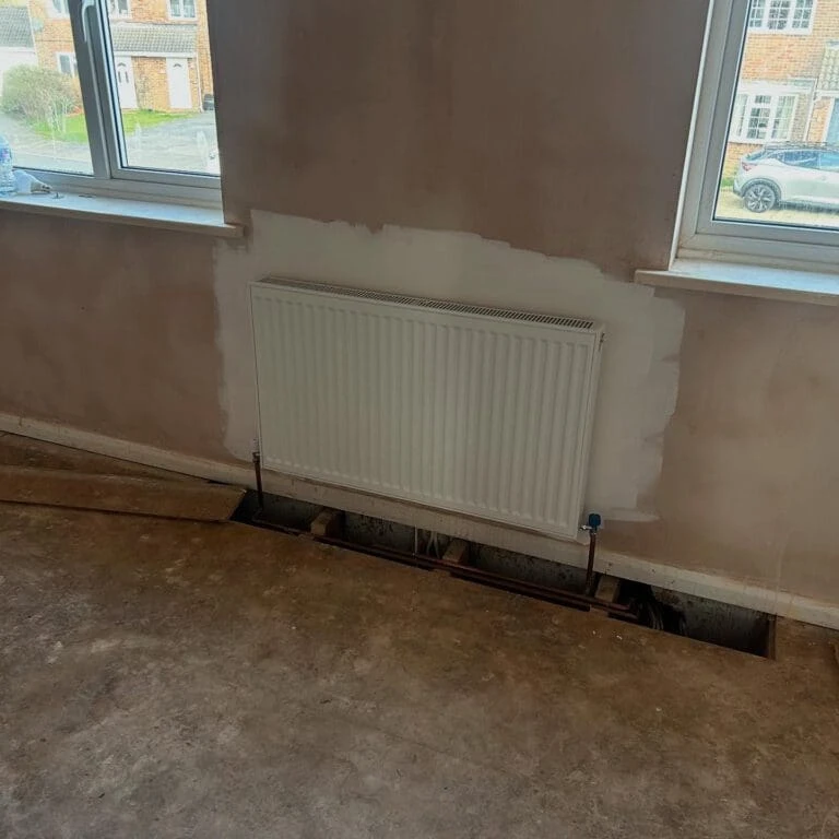 Interior view of a room with a radiator on a wall and partially finished flooring, revealing exposed pipes underneath.