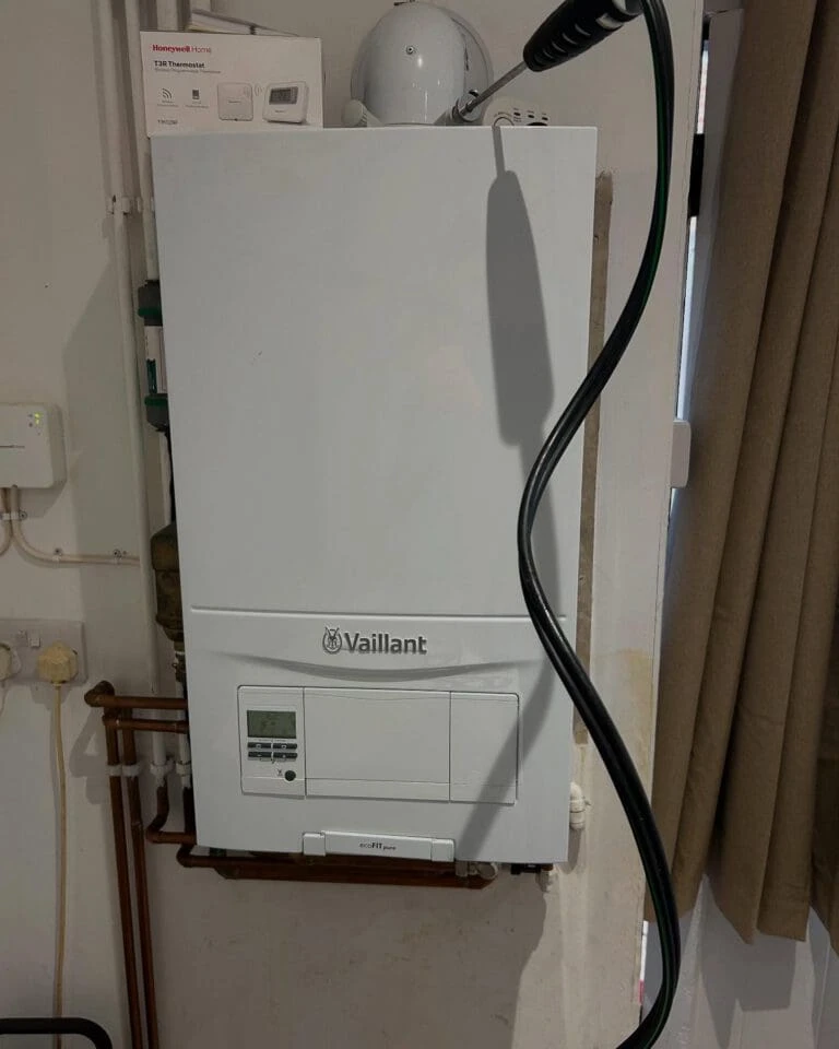 A white Vaillant boiler mounted on a wall, with a digital display and pipes below. A green hose is coiled beside it.