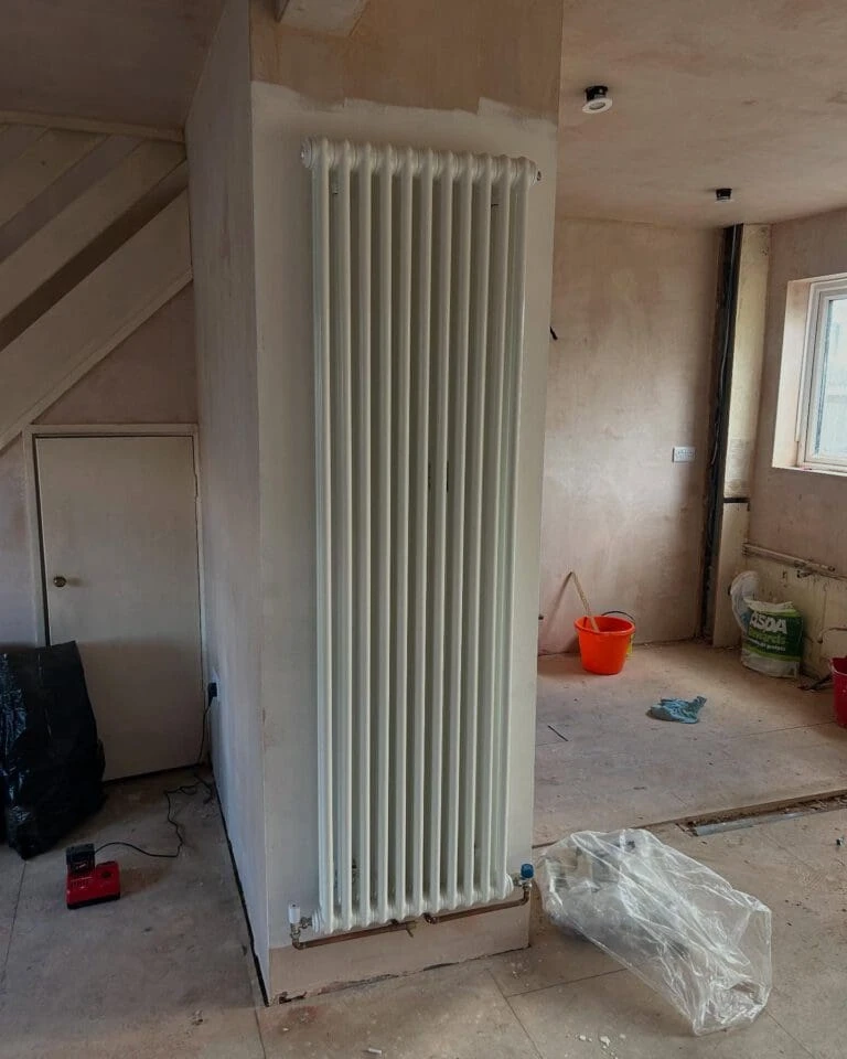A tall, white radiator is installed on an unfinished wall in a room with bare walls and a wooden staircase nearby. Various tools and a bucket are visible.
