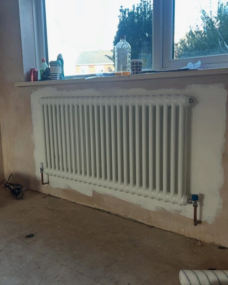 A white radiator mounted on a wall, with tools and containers on the windowsill in a partially renovated room.