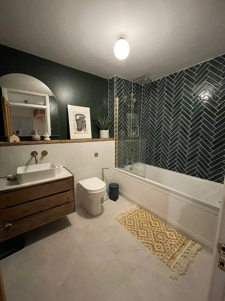 Modern bathroom with dark green walls, herringbone tile, a bathtub, wooden sink cabinet, and decorative rug. Good lighting and plants.