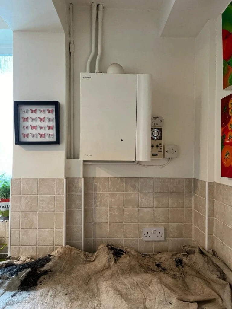 A kitchen corner featuring a wall-mounted boiler, electrical outlets, tiled backsplash, and a butterfly artwork in a frame.