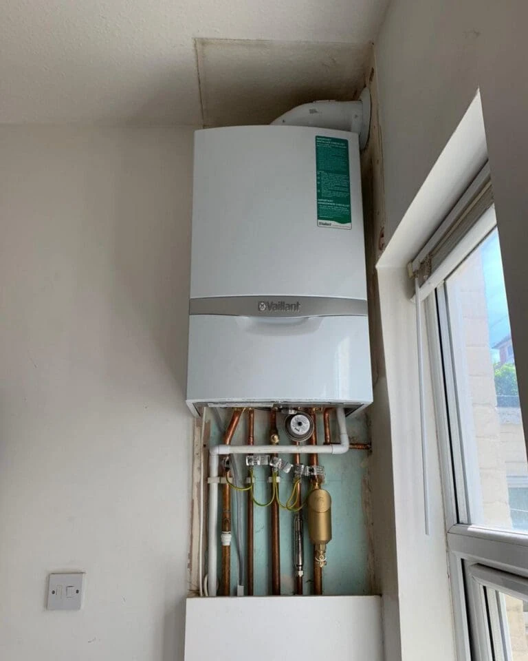 A white boiler mounted on a wall, showing connected pipes and a control panel, beside a window.