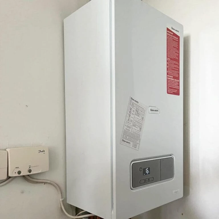 A white wall-mounted boiler with a digital display and instructions attached, next to a small control panel on the wall.