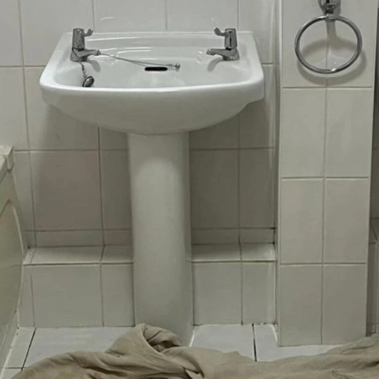 A small white sink with two faucets, positioned next to a toilet, on tiled bathroom floor with a beige cloth nearby.