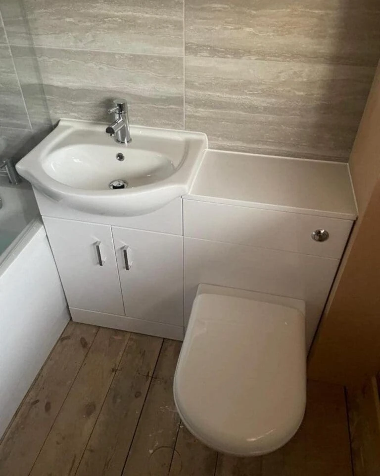 Compact bathroom setup featuring a white sink and toilet against a gray tiled wall, with wooden flooring.