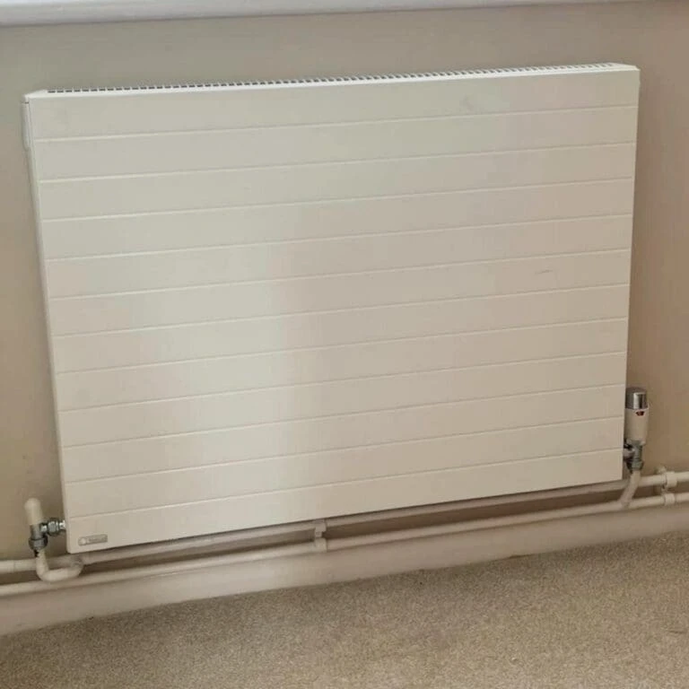 A white horizontal radiator mounted on a wall, with visible pipes below. The background is plain and light-colored.