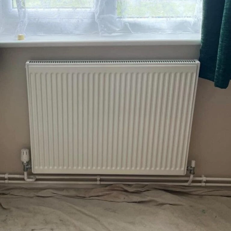 A white vertical radiator installed under a window, with visible piping and a curtain to the right. The floor is covered with a protective sheet.