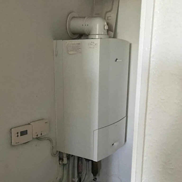 A white boiler installed in a corner, featuring a vent pipe and connected controls nearby, mounted against a light-colored wall.