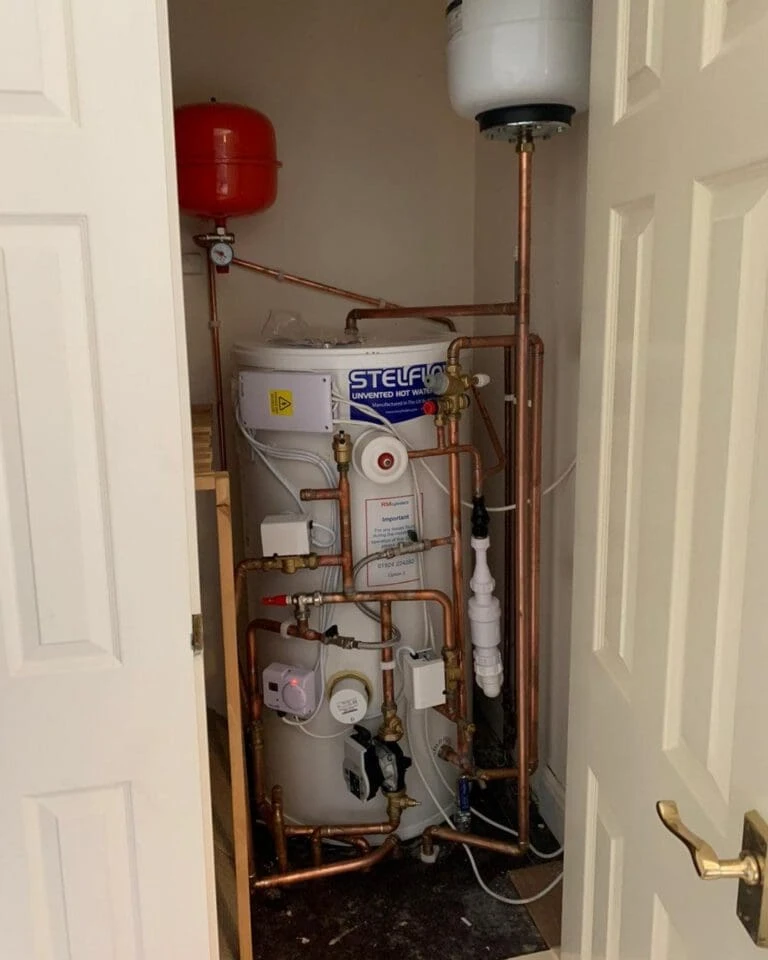 A hot water tank with copper pipes and red pressure vessel is installed in a corner behind a door.