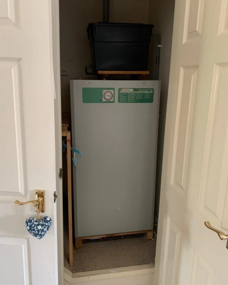 A door opens to a small closet containing a gray appliance with a pressure gauge, a black storage container on top, and ambient lighting.