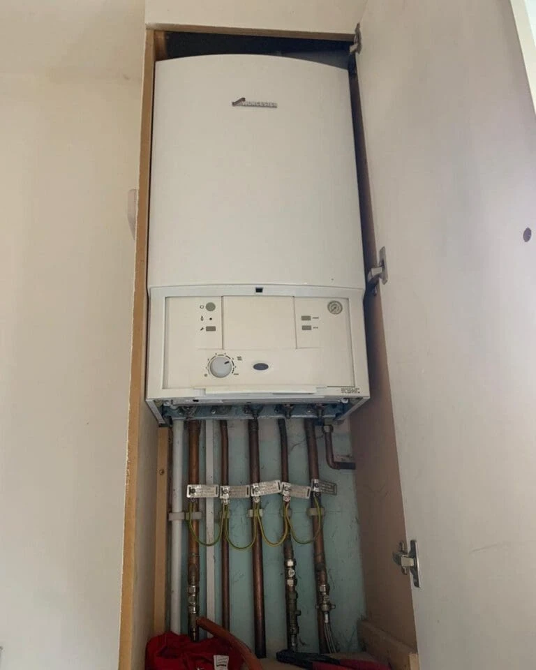 A white boiler is installed inside a closet. Below it are metal pipes connected to valves, with a red bag partially visible at the bottom.