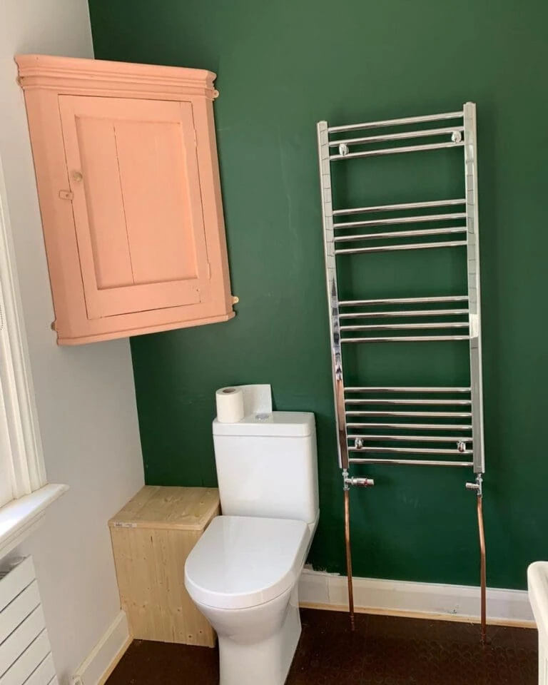 Modern bathroom with green walls, a pink cabinet, a white toilet, wooden storage, and a chrome towel warmer mounted on the wall.