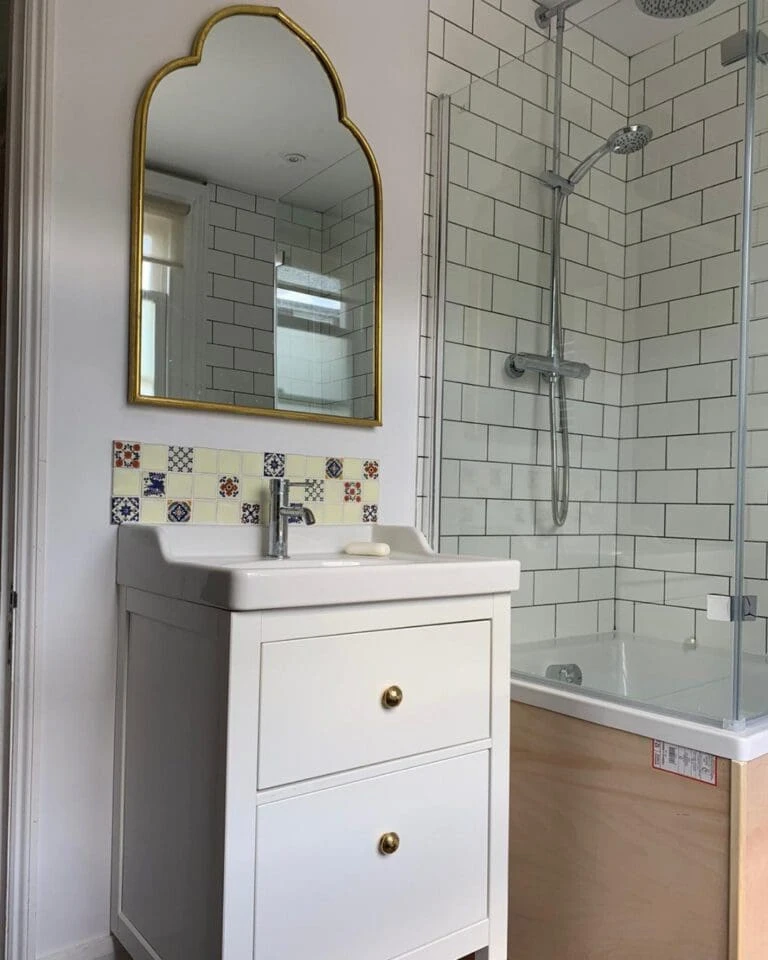 Bright bathroom featuring a white vanity with gold hardware, a large arched mirror, and tiled shower with glass doors.