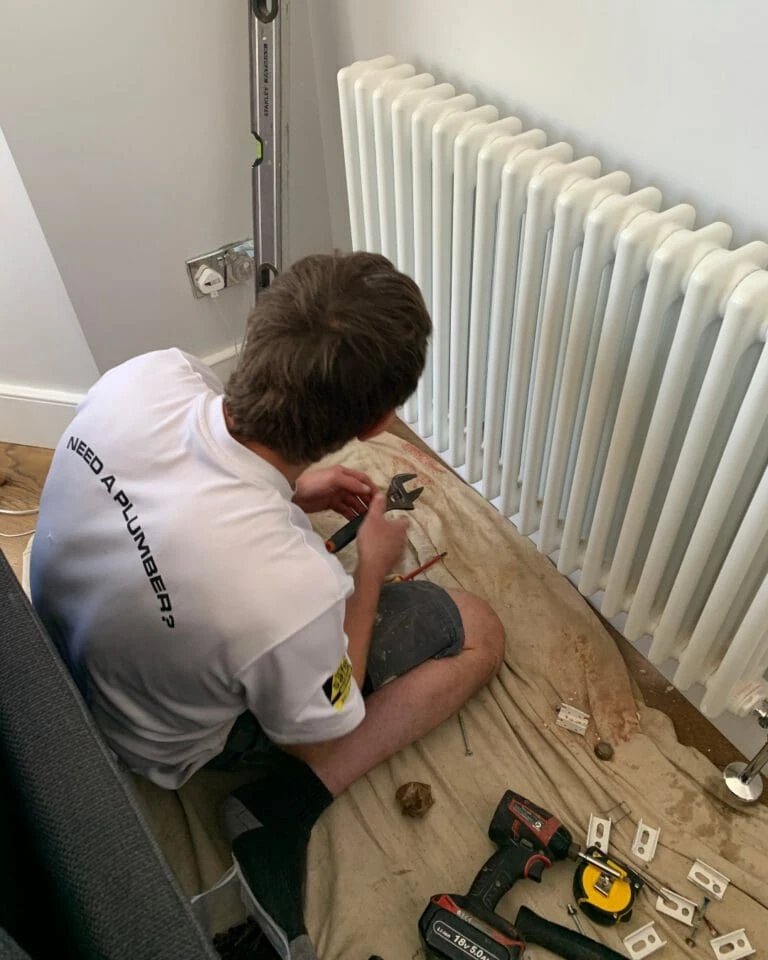 A person kneels on a cloth, using pliers to work on plumbing near a white radiator, with tools scattered around.
